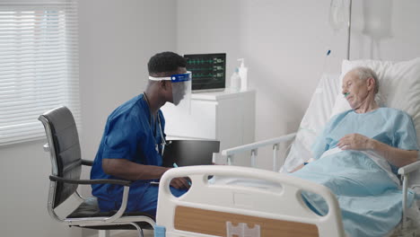 a black male doctor in a protective mask comes in and talks to patients with an elderly white man lying on a hospital bed