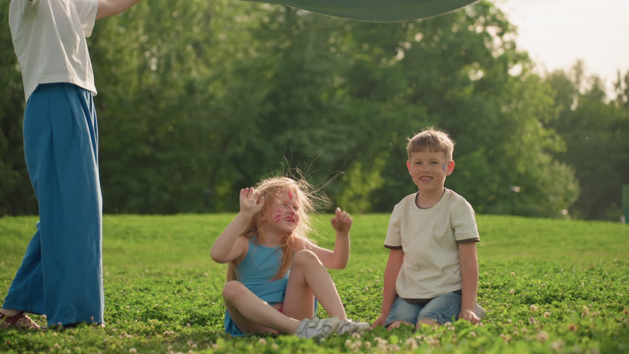Joyful moms holding two edges of cloth while kids in center laugh and play as fabric swings over them in sunny park, green grass background, casual summer clothes, family bonding moment