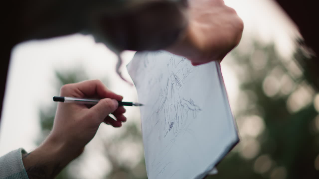 Caucasian hiker sketching trail map outdoors closeup of tattooed hands holding paper pad and pen, sketching topographic map lines, blurred pine forest background, soft sunset light, candid travel