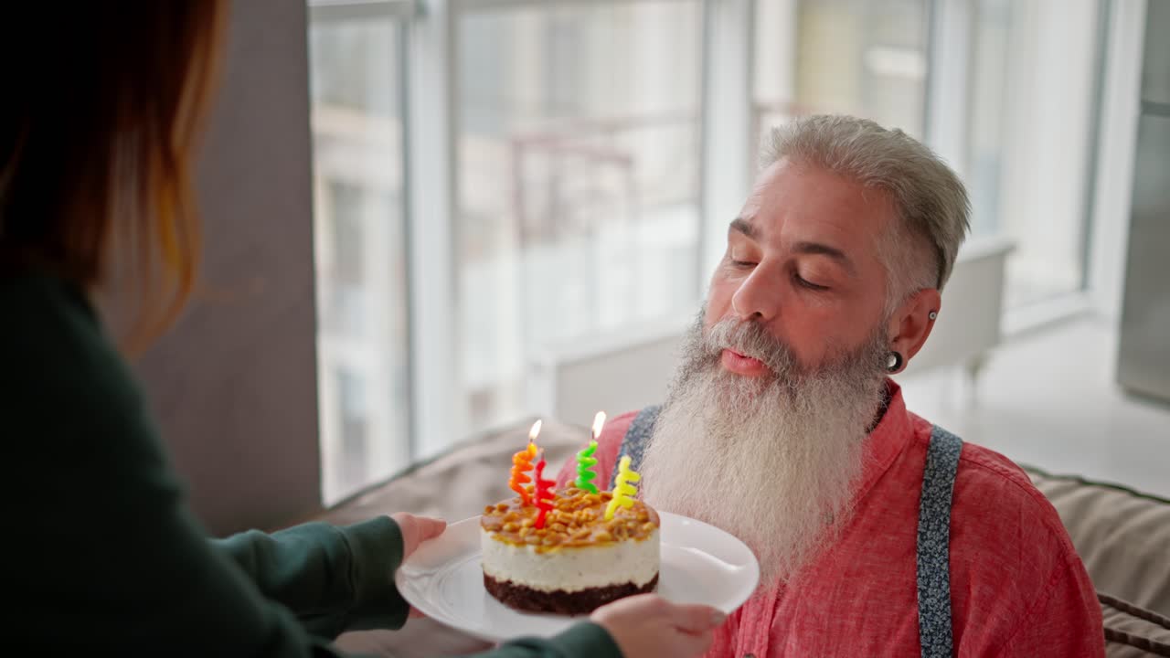 un hombre feliz con cabello gris y una barba exuberante en una camisa rosa sopla cuatro velas y en un pequeño pastel durante su cumpleaños y felicitaciones de su hija adulta en un apartamento moderno