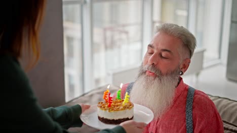 Ein-Glücklicher-Mann-Mit-Grauem-Haar-Und-üppigem-Bart-In-Einem-Rosa-Hemd-Bläst-Vier-Kerzen-Aus-Und-Auf-Einem-Kleinen-Kuchen-An-Seinem-Geburtstag-Und-Glückwünsche-Von-Seiner-Erwachsenen-Tochter-In-Einer-Modernen-Wohnung