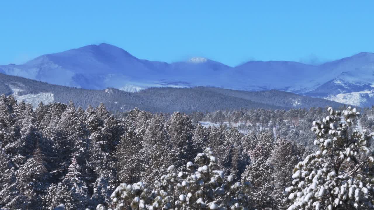Pemandangan udara drone di musim dingin Colorado: Gunung Blue Sky, Pegunungan Rocky Front Range Gunung Evans. Pagi hari dengan bayangan awan, salju, dan langit biru cerah. Terlihat konifer malar hijau, Denver, puncak berselimut salju yang menonjol, serta pepohonan hutan bersalju. Bidikan dengan perbesaran.