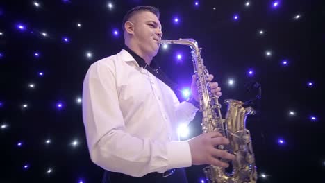 tilt up shot of biracial jazz musician in checkered suit and bowtie playing saxophone while sitting against black band member in dimly lit bar, rehearsing and checking sound before live performance
