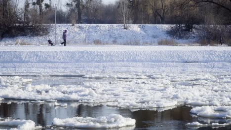 Frau,-Die-Kind-Auf-Schlitten-Neben-Dem-Fluss-Mit-Eisschollen-Zieht,-Statischer-Schuss