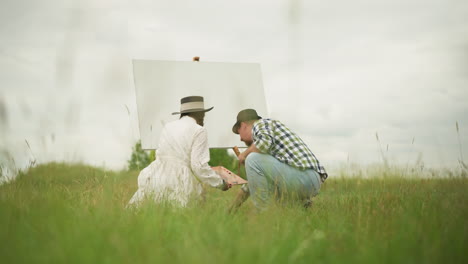 two individuals bent over in a grassy field, focused on their tasks. the woman in a white dress holds a palette, while the man in a checked shirt and hat reaches for something on the ground