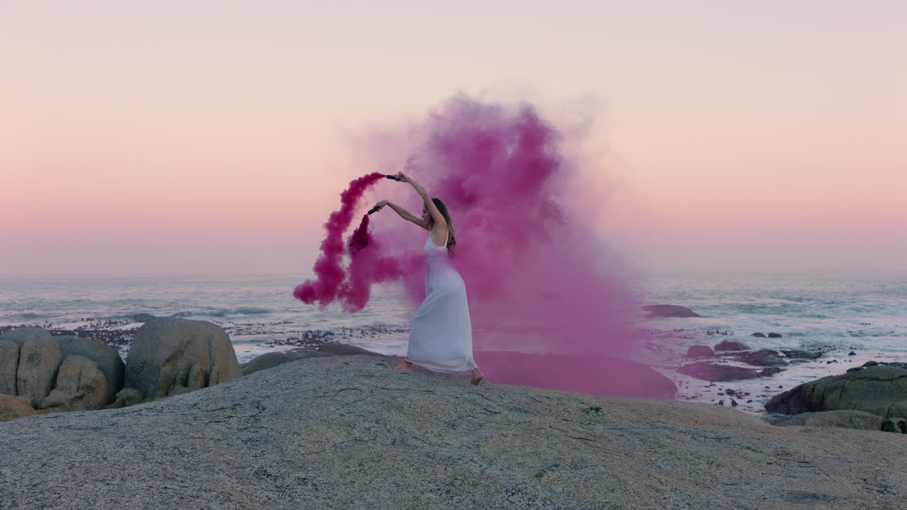 핑크색 연기 폭탄을 들고 있는 여성이 이른 아침에 해변에서 춤을 추고 창조적 자유를 축하하고 있습니다.