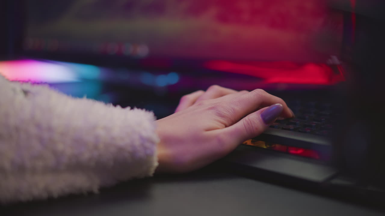 Hand view of artist with polished nails resting on illuminated mechanical keyboard during gaming session, with partial view of vibrant screen and colorful ambient lighting