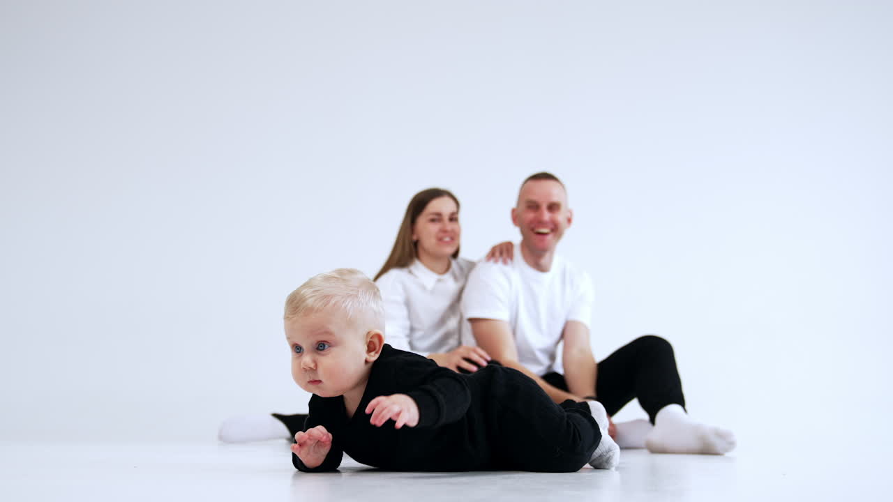Positive Caucasian parents send air kiss to their baby sitting in front of them. Little male kid crawls by the floor. White backdrop.