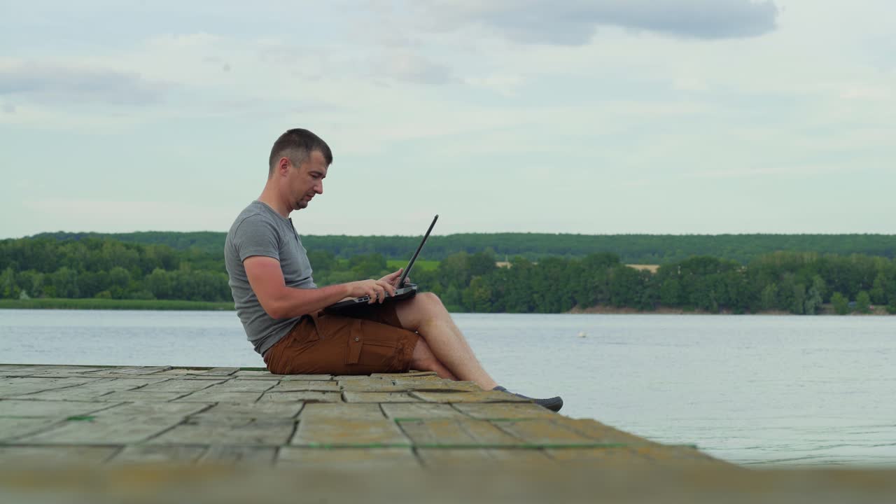 man is sitting on the bridge and opening a laptop to work on the background of the river in the summer
