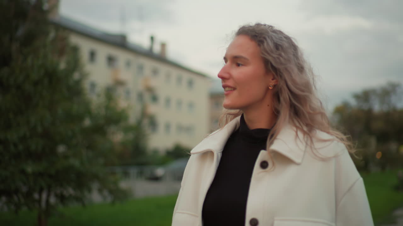 Freelancer twirling closed umbrella while admiring distant view, wearing light coat with soft smile, walking outdoors with blurred office building, green trees, and urban elements