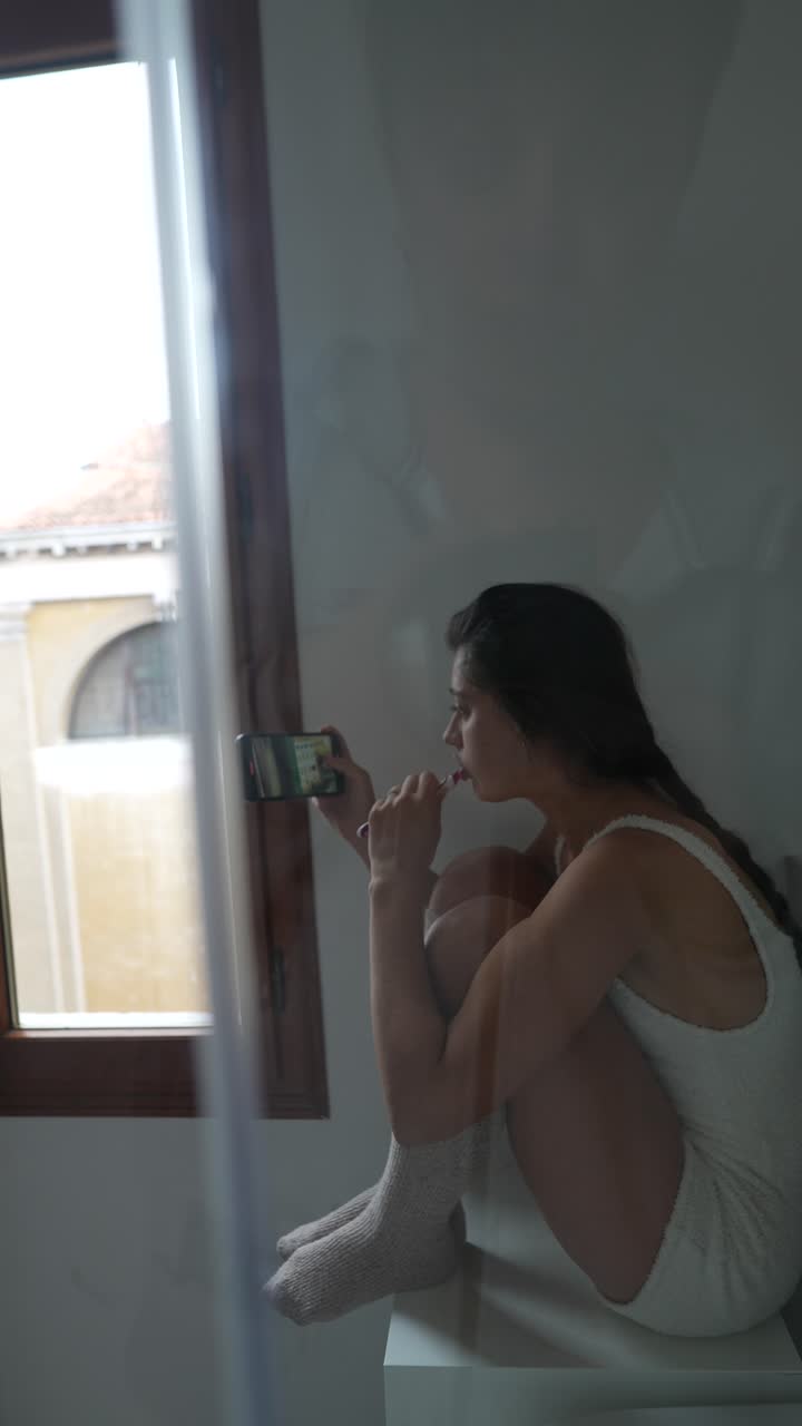 Woman brushing teeth while using phone in a bathroom near a window