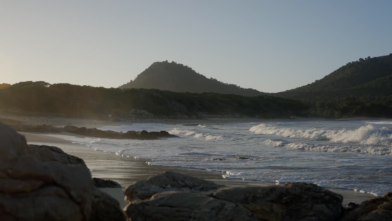 Onde oceaniche che si infrangono sulla spiaggia al tramonto con montagne sullo sfondo.