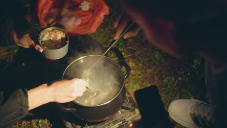 close up of people cooking outdoors as one stirs steaming pot while other turns canned food into pot, man uses phone light for visibility during nighttime camping meal preparation on forest ground