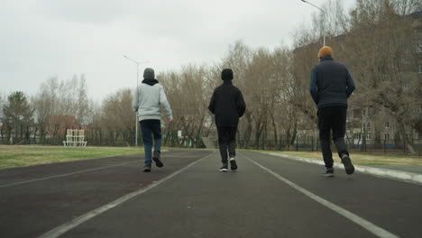 back view of an adult and two young boys jogging side by side on a track in a stadium, with a residential building visible across the road, with bare trees on the right hand side