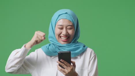 close up of asian muslim woman looking at smartphone then screaming goal celebrating while standing in the green screen background studio