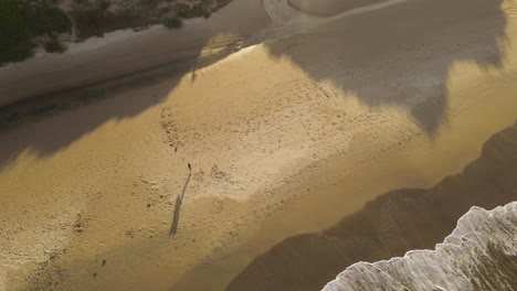 Toma-Aérea-De-Arriba-Hacia-Abajo-De-Un-Joven-Surfista-Que-Sale-Del-Agua-Caminando-En-Una-Playa-De-Arena-Al-Atardecer