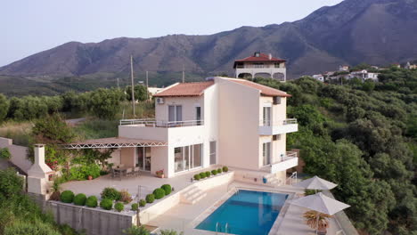 vuelo aéreo en sentido antihorario de una lujosa villa griega con piscina con montañas al fondo y follaje verde en primer plano