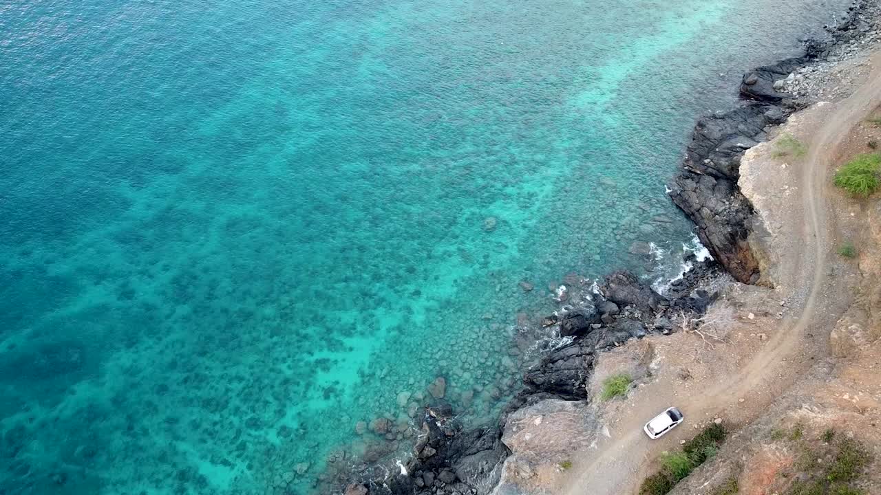 un coche blanco viaja lentamente por una carretera costera sinuosa, accidentada y llena de baches con un hermoso océano de aguas turquesas