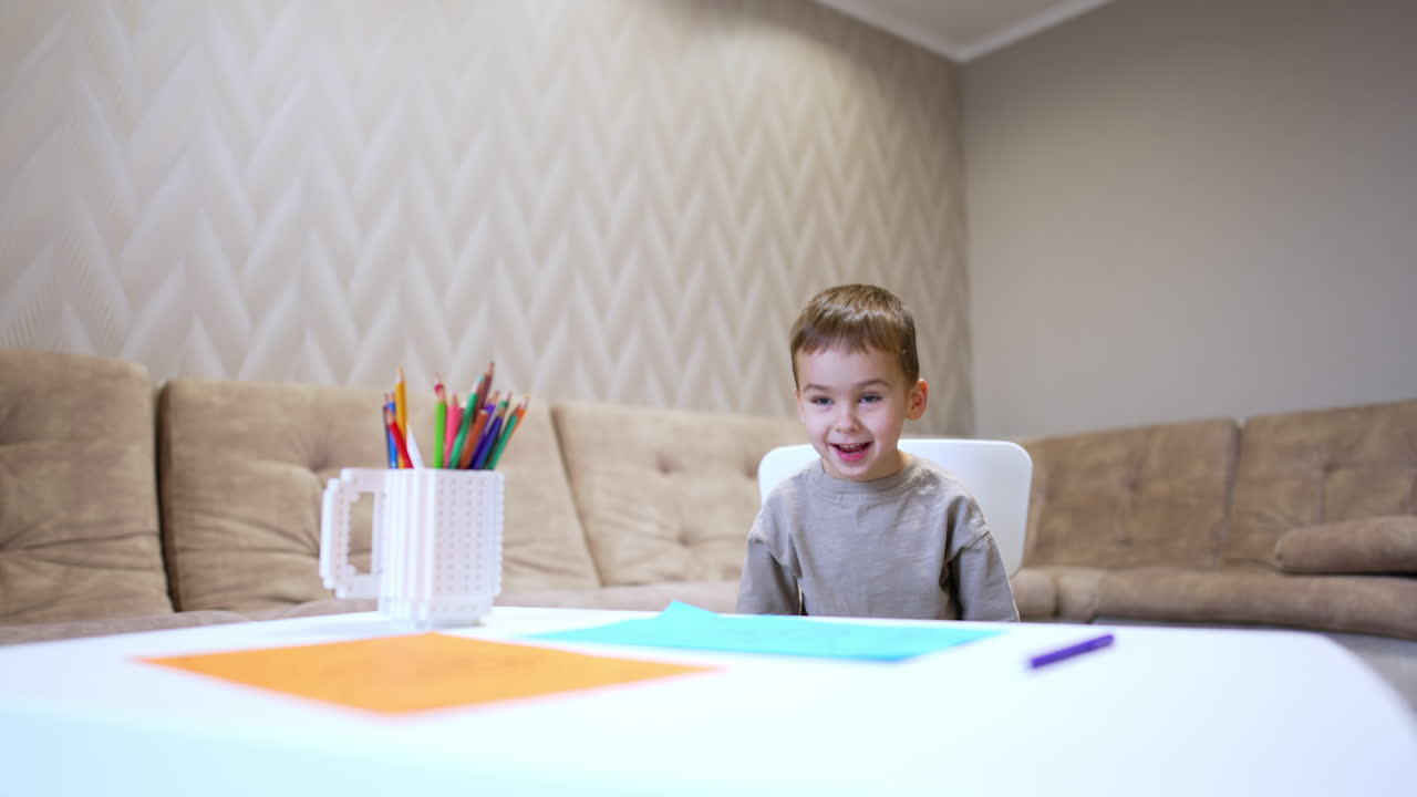 Small cute baby boy standing at the desk sits down on the chair. Kid moves away from the desk on his chair.