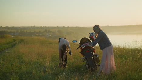 two ladies are seen in an open grassy field near a motorcycle, one is bent over with hands in a bucket, while the other stands cleaning the bike s back seat, close to a river bank