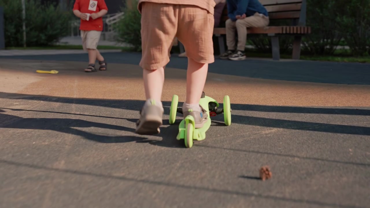 Young boy rides green kick scooter on outdoor path with other people visible in background. scene captures his movement through sunlight and shadow details emphasizing active play in public