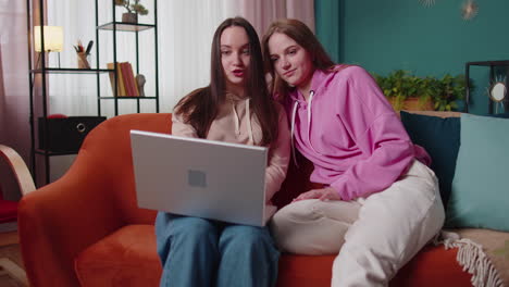 happy two female women couple family on sofa at home, enjoy chatting, spend leisure time on laptop