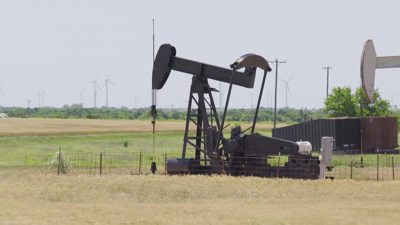 Drone 4K of a wheat field with two oil pump jacks and wind turbines in the background.