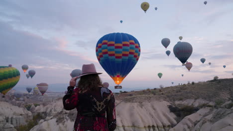 female tourist in long dress and hat watching colorful hot air balloons flying in the sky at sunrise in cappadocia, turkey