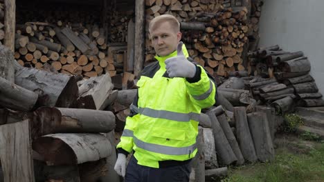 Lumberjack-in-reflective-jacket.-Man-woodcutter-show-thumb-up.-Sawn-logs,-firewood-background