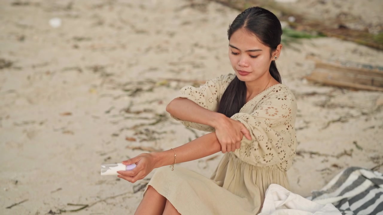 Asian woman applying sunscreen on beach, squeezing lotion from bottle and smoothing cream over arms, braided hair with flower, seated on striped towel near driftwood, sunsafe summer skincare routine