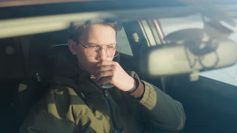 close up through windshield of young man in hoodie and sunglasses eating pastry while sipping coffee from travel mug seated in parked car with blurred winter city background