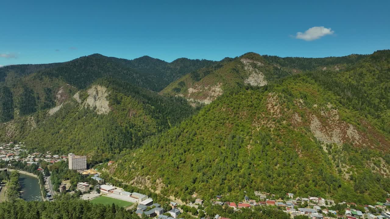 A wide aerial view of a valley town nestled beneath rocky mountain slopes, surrounded by dense forest and bright midday light