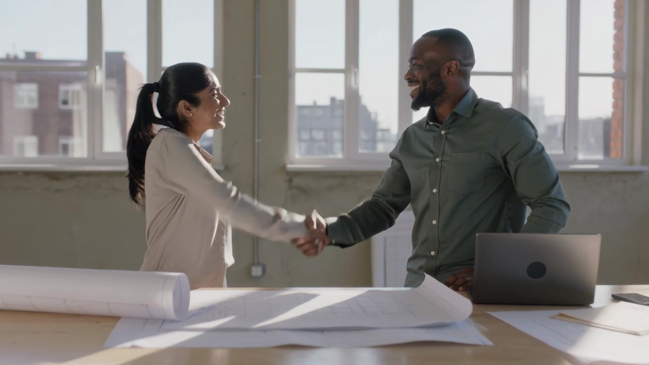 Architects in shirts rising, shaking hands at loft table after brief exchange over plans and laptop