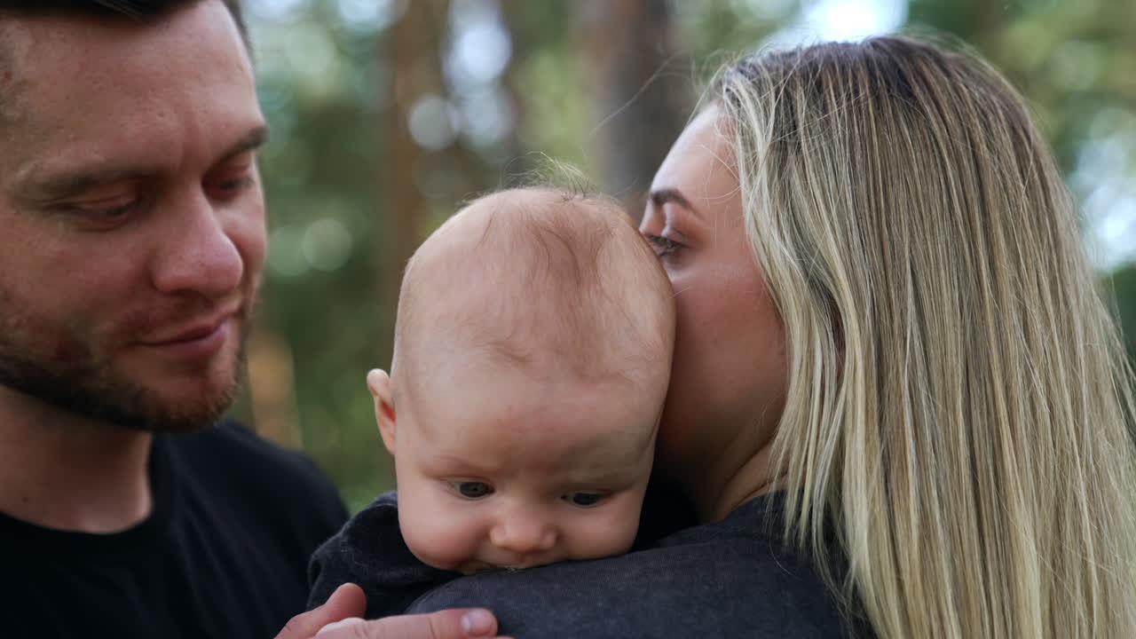 Adorable baby leaned to his mom's shoulder. Bearded man kisses his son on the head. Close up. Blurred backdrop.