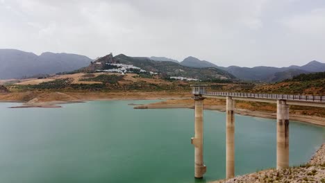 Smooth-aerial-lateral-scene-of-a-dam-machinery-a-bluish-lake-and-a-medieval-castle-on-the-top-of-a-mountain