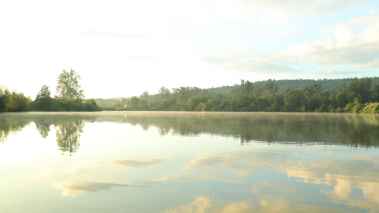 아침 안개가 표면에 반사된 떠오르는 태양과 함께 물 위로 돌진하는 호수의 잔잔한 수위의 전망과 호수의 잔잔한 표면 뒤에 멀리 물고기가 나타납니다