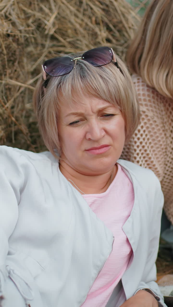 중년 여성은 개 줄을 들고 누워서 사려 깊게 보이지만, 그녀의 친구는 멀리서 쳐다보고, 열린 시골에서 풀 근처에서 편안한 순간을 보내고 있습니다.