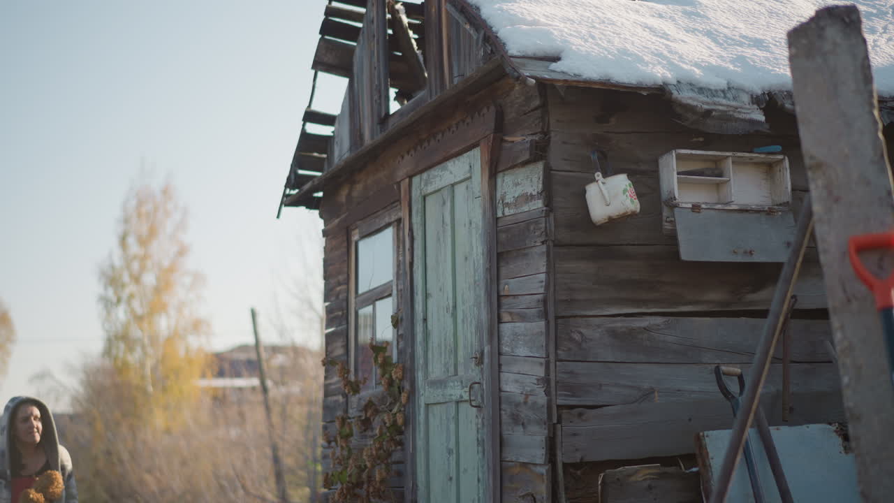 Tetto innevato di una casa rustica con donna che si avvicina tenendo un bouquet essiccato, pala appoggiata alla porta, assi stagionate e camino, pallida luce mattutina, brina che si scioglie sulle tegole, resilienza domestica