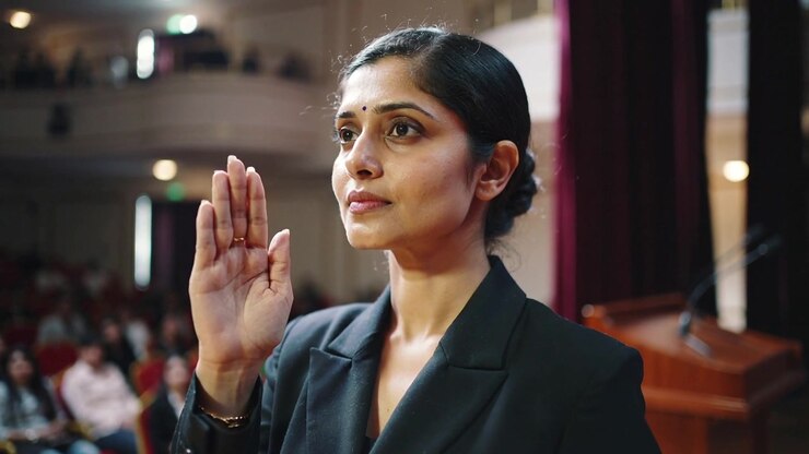 Holding raised right hand woman in dark blazer with name badge near lectern during oath taking