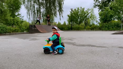 cheerful little boy jumping up sitting on his toy car outdoors. toddler watching the older boys ride their scooters at backdrop.