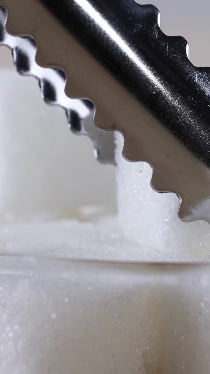 Close-up of silver tongs lifting a refined sugar cube from a clear glass bowl.