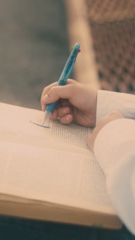 young guy in white shirt and glasses draws in paper notebook with blue pen sitting on brown bench close view