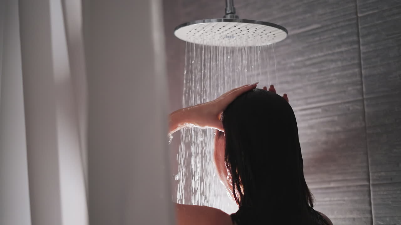 갈색 여성은 샤워에서 머리카락과 몸을 습니다. 여성은 회색 타일로 현대적인 샤워장에서 머리카락을 니다. 큰 샤워실의 물은 여성의 몸 아래로 흐릅니다.