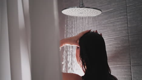 mujer morena lava el cabello y el cuerpo en la ducha. dama acaricia el cabello en la ducha moderna con azulejos grises. agua de la ducha grande fluye por el cuerpo de la dama