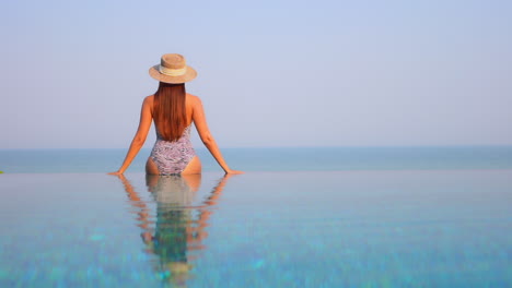 A-sexy,-healthy-young-woman-in-a-zebra-bathing-suit-and-a-straw-hat-sits-on-the-invisible-edge-of-a-resort-pool-to-look-out-at-the-ocean-horizon-beyond