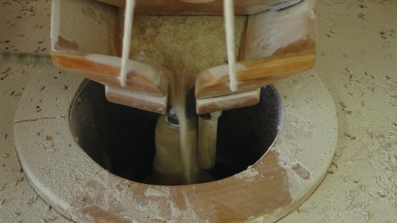 Close-up de um moinho de farinha tradicional em funcionamento, com grãos sendo moídos em farinha através de um mecanismo de madeira e poeira fina ao redor da área de moagem.