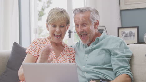 Senior-Retired-Couple-Sitting-On-Sofa-At-Home-Making-Video-Call-On-Laptop