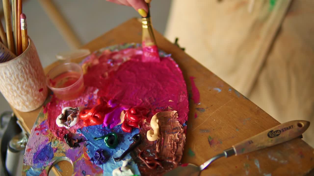 artista femenina con pincel grueso está mezclando diferentes pinturas rosadas de aceite de colores en la paleta. vista de cerca de la mano femenina mezclando colores. paleta colorida. ángulo alto