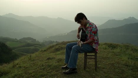 asian musician sits on countryside hills playing guitar music in vintage clothes male performer in vintage clothes and sunglasses