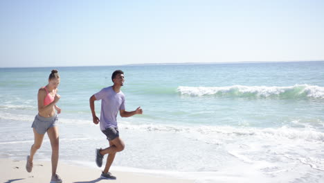 una joven caucásica y un hombre biracial están corriendo en una playa soleada con espacio para copiar